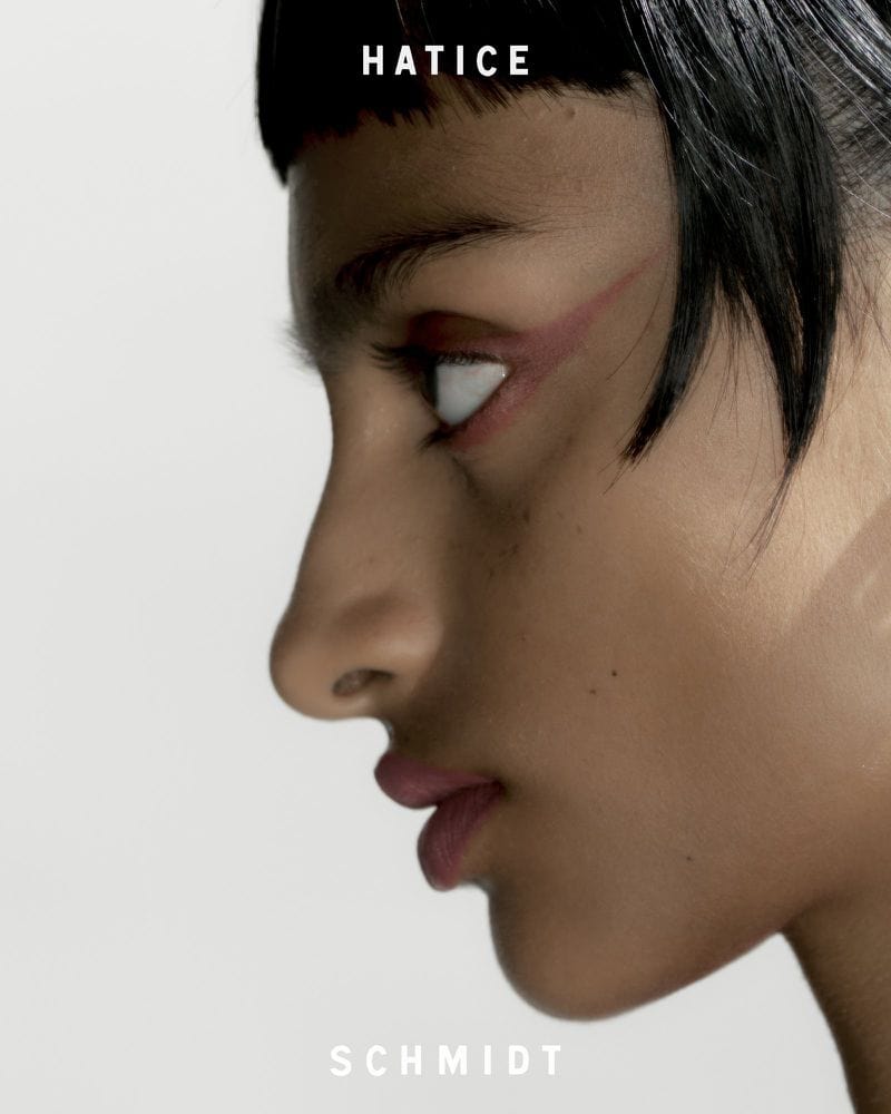 Close-up profile portrait of a model with dark fringe hair, bold eye makeup and neutral lips against a plain white background.