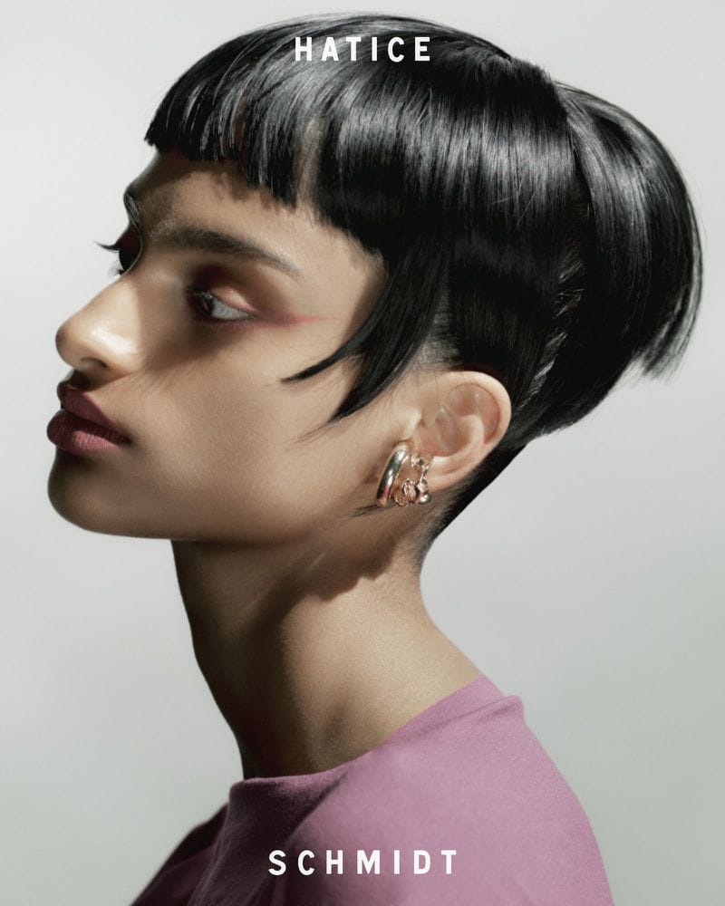 Profile portrait of a woman with dark hair and pink lipstick wearing metallic ear cuffs against a light grey background