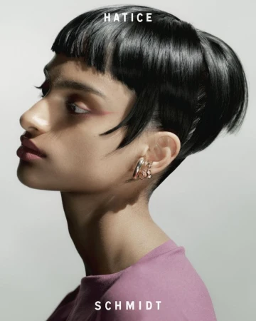Profile portrait of a woman with dark hair and pink lipstick wearing metallic ear cuffs against a light grey background