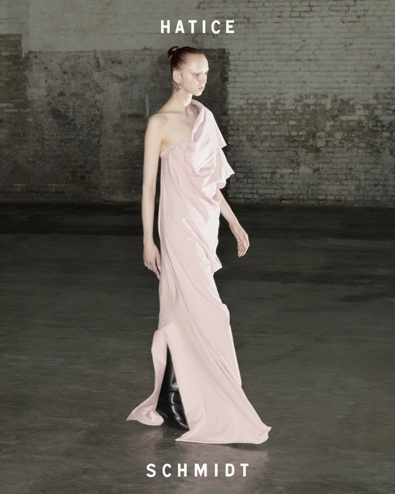 Model in a pale pink one-shoulder gown walking against a distressed brick background in a campaign by Hatice Schmidt.