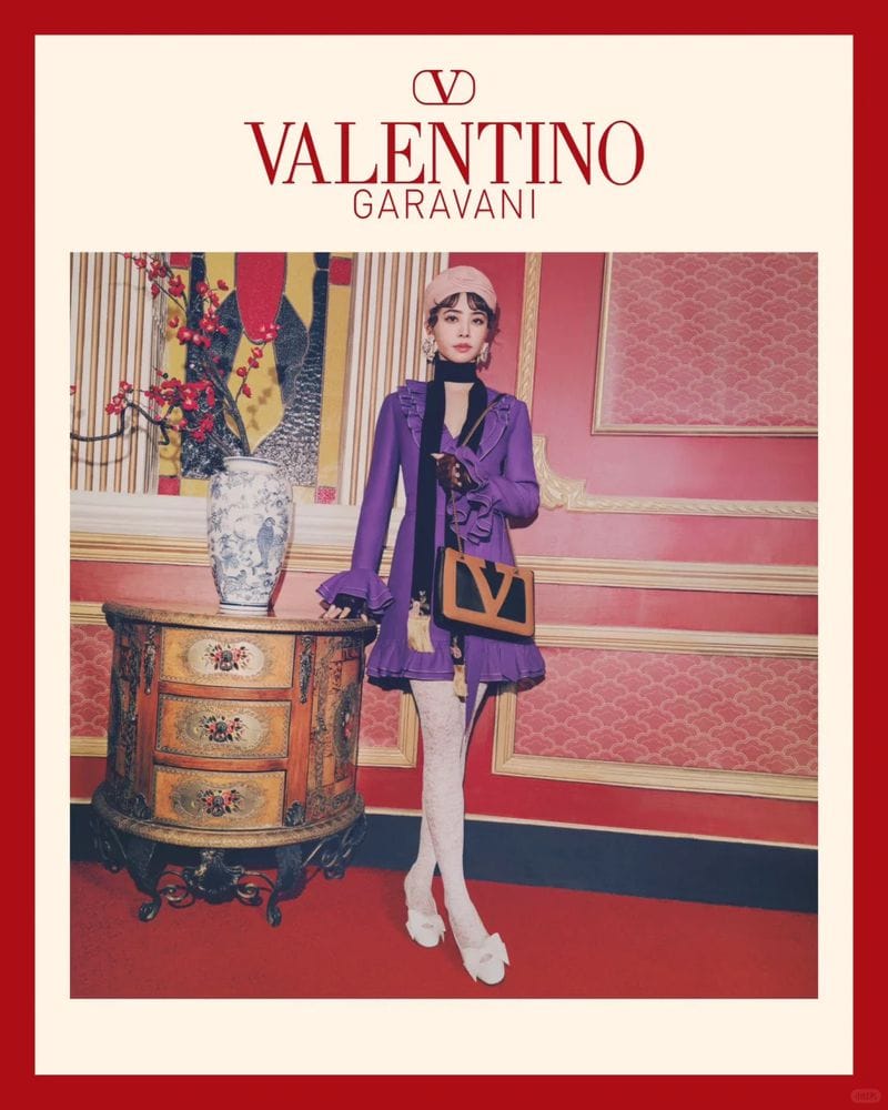 Woman in a purple dress and beret holding a structured handbag in front of a red-paneled wall with decorative furniture
