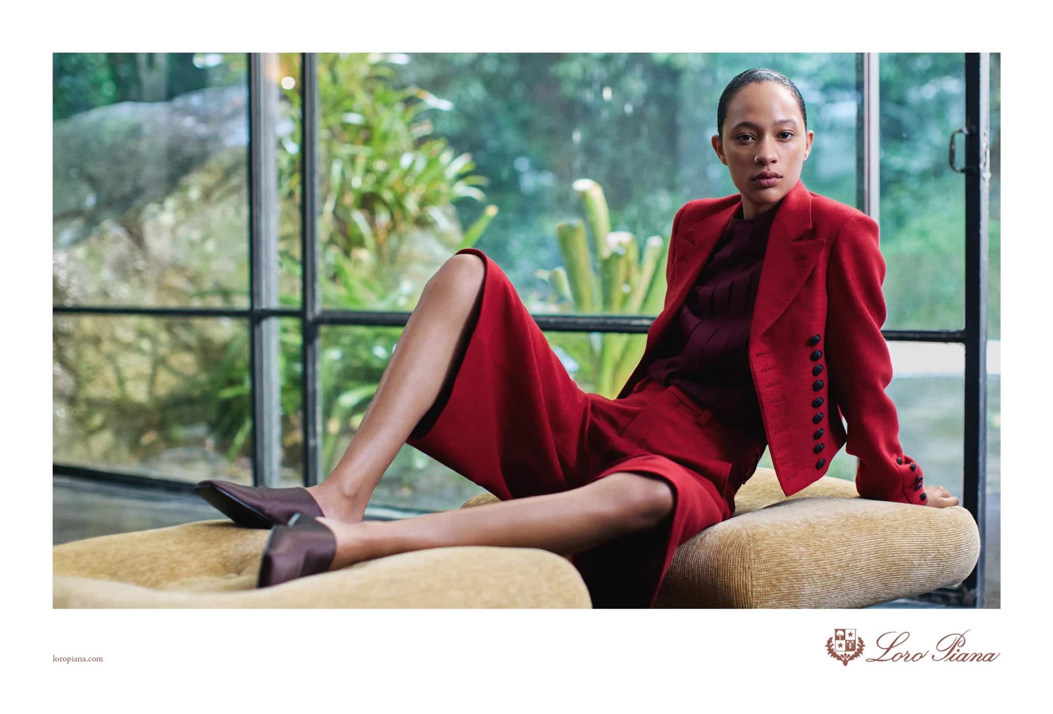 Model wearing a red blazer and shorts seated indoors against large windows with lush green foliage outside