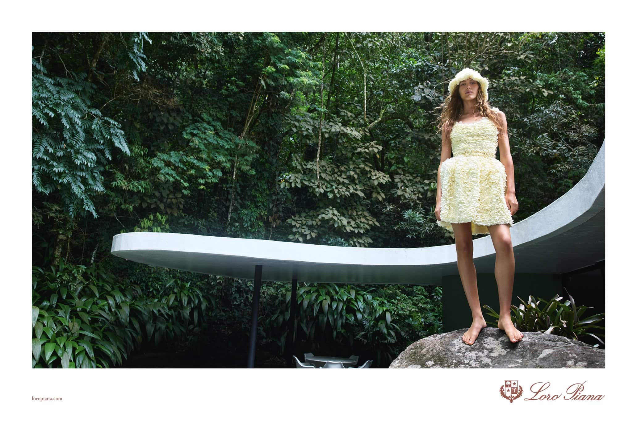 Model standing barefoot on a rock wearing a textured yellow dress and matching hat outdoors amidst lush green foliage.