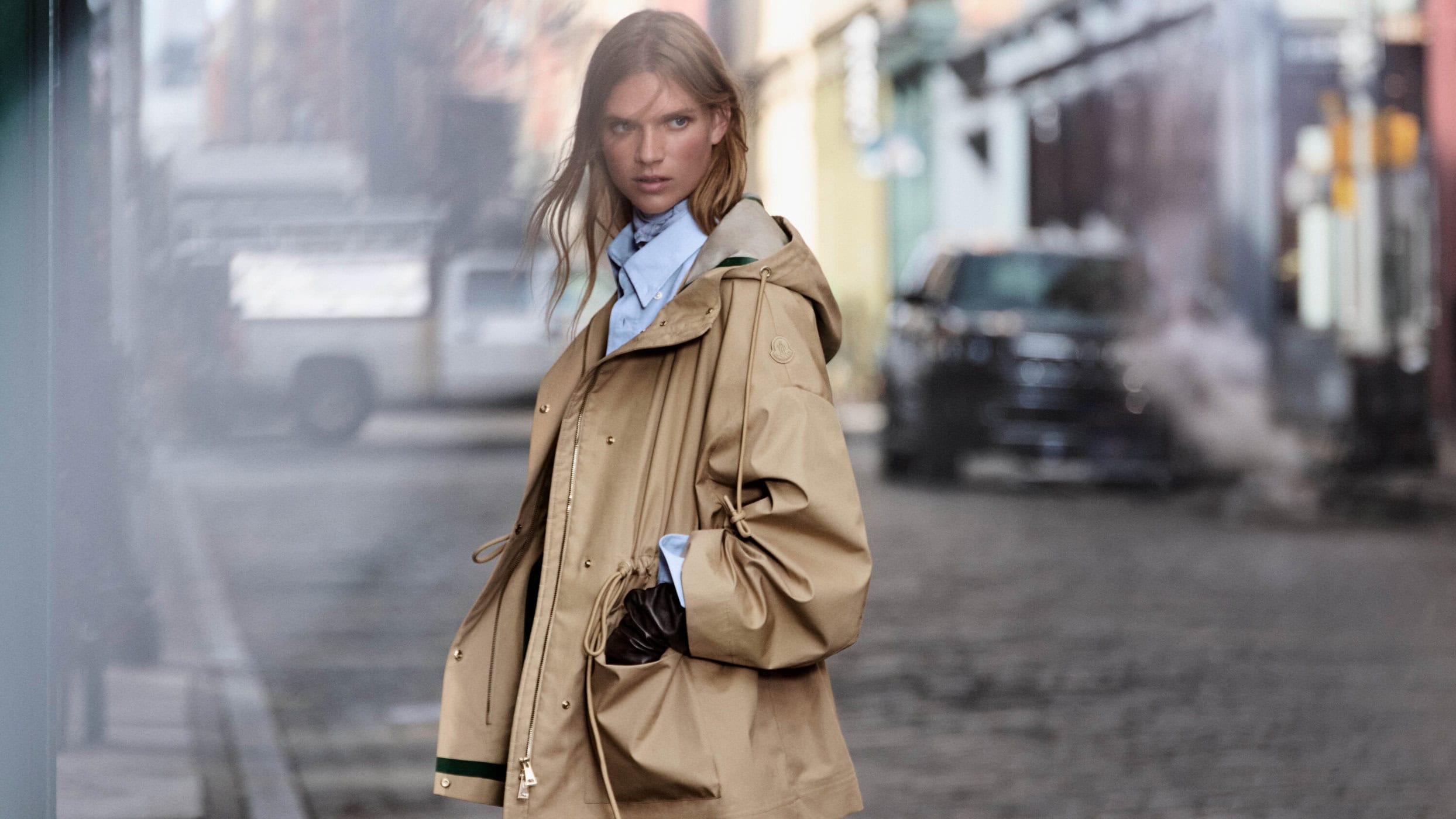 Model in a tan hooded parka and light blue shirt standing on a city street for Moncler Spring 2025 campaign