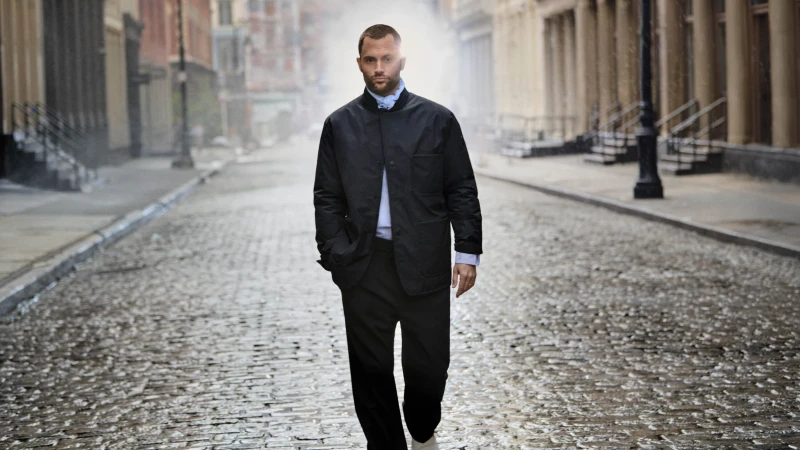 Penn Badgley walking on a cobblestone street wearing a black jacket and dark trousers for Moncler