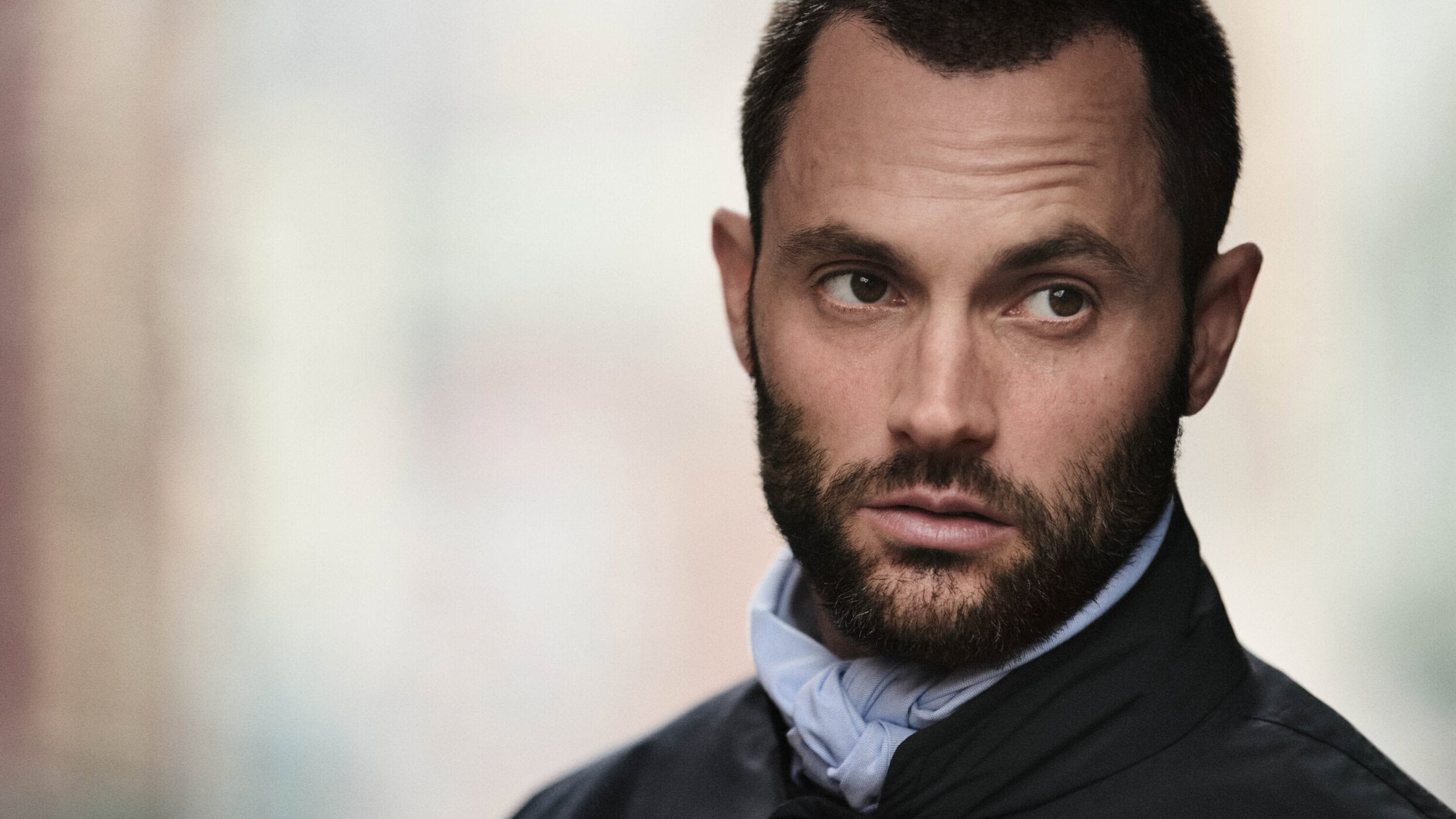 Portrait of Penn Badgley with short dark hair and a beard wearing a dark jacket and pale blue neck scarf against a soft blurred background