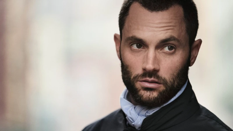 Portrait of Penn Badgley with short dark hair and a beard wearing a dark jacket and pale blue neck scarf against a soft blurred background