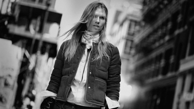 Model in quilted puffer jacket and button-down shirt walking outdoors on a city street in a black and white advertising campaign image.