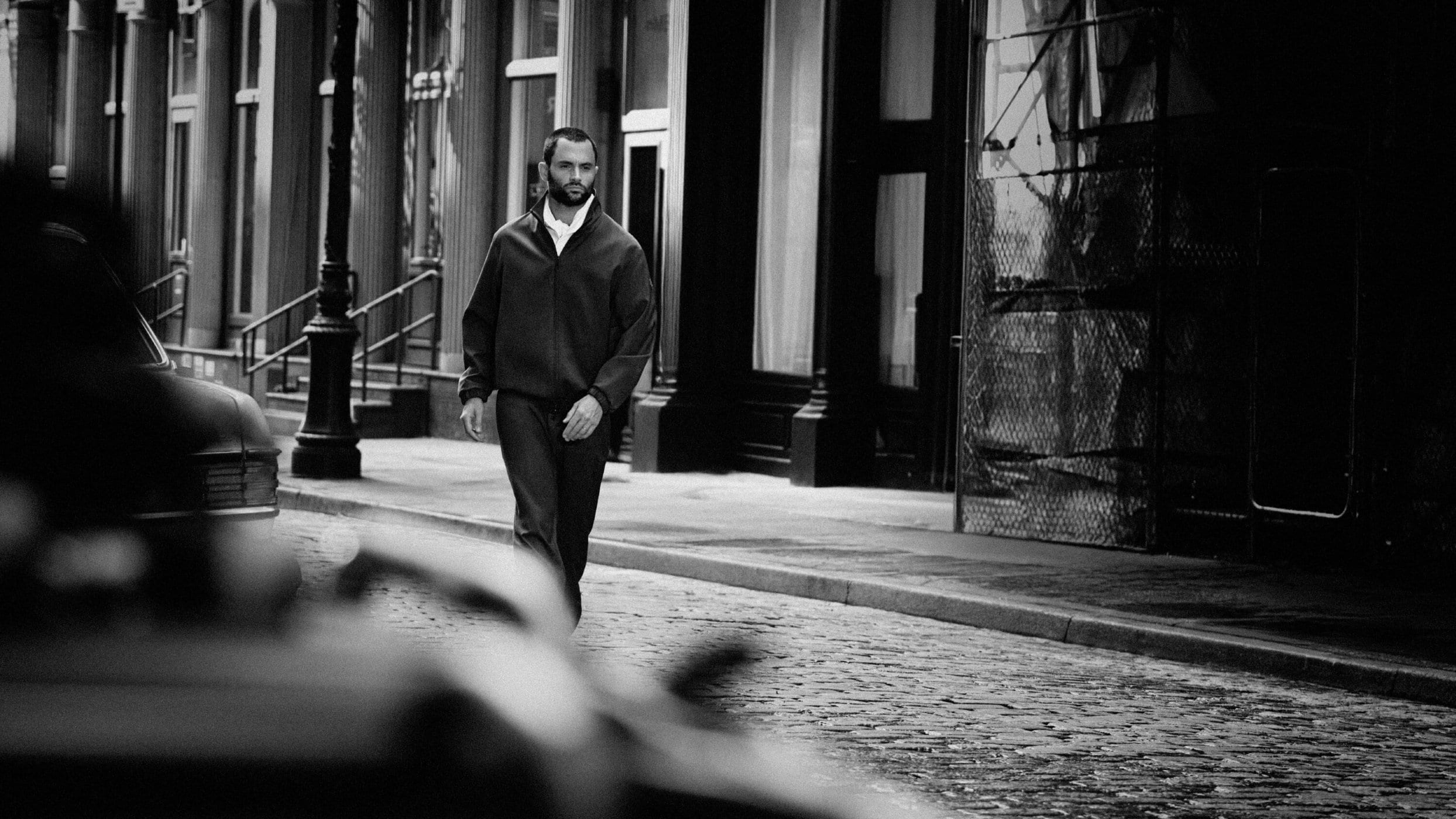 Man walking on a cobblestone street wearing a dark zip-up jacket in a black and white campaign image.