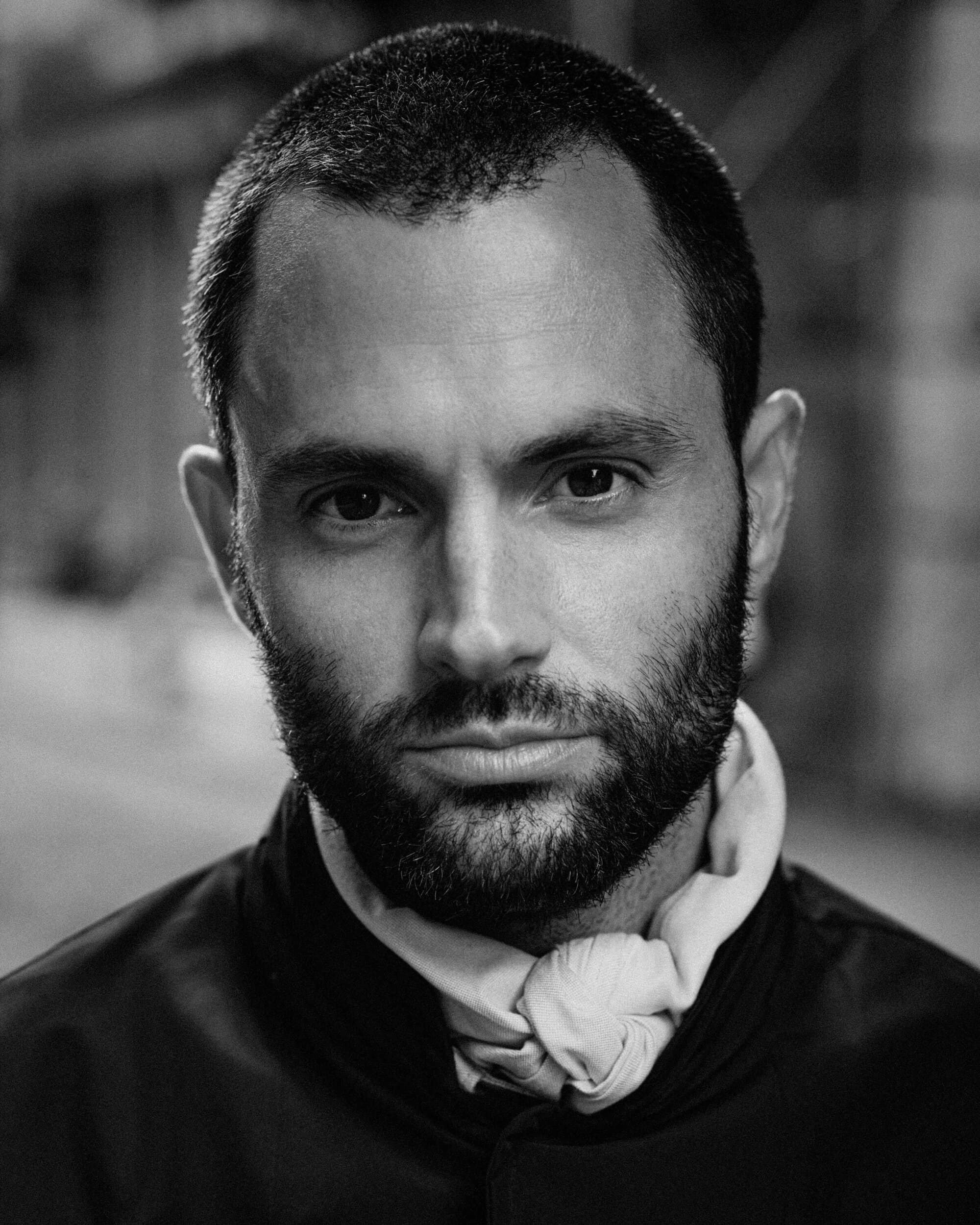 Close-up black and white portrait of Penn Badgley wearing a dark jacket and knotted scarf for Moncler spring 2025 ad campaign.