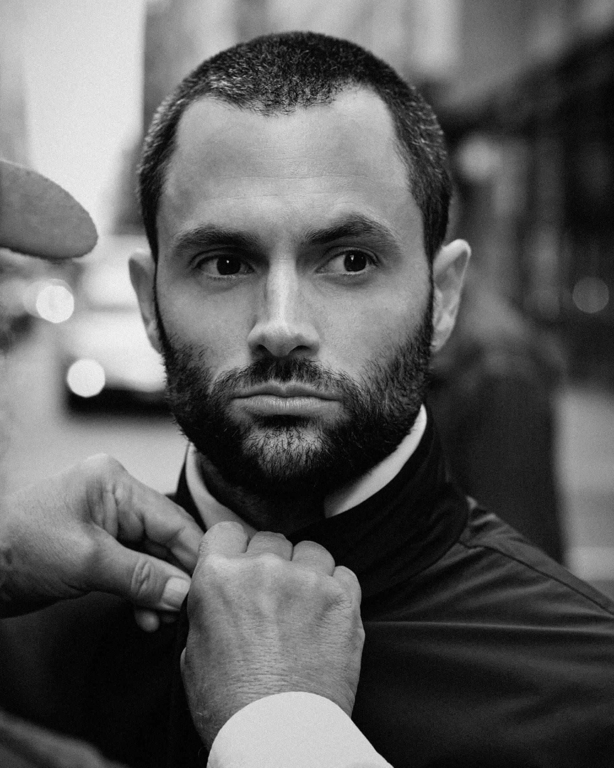 Close-up black and white portrait of Penn Badgley getting his jacket collar adjusted for Moncler.