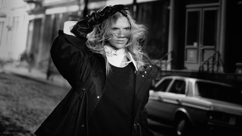 Monochrome fashion campaign image featuring a model wearing a dark jacket and leather gloves on an urban street.