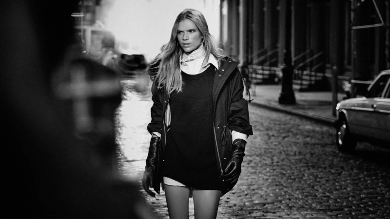 Monochrome street-style campaign image featuring a model walking on a cobblestone road wearing a dark parka and gloves.