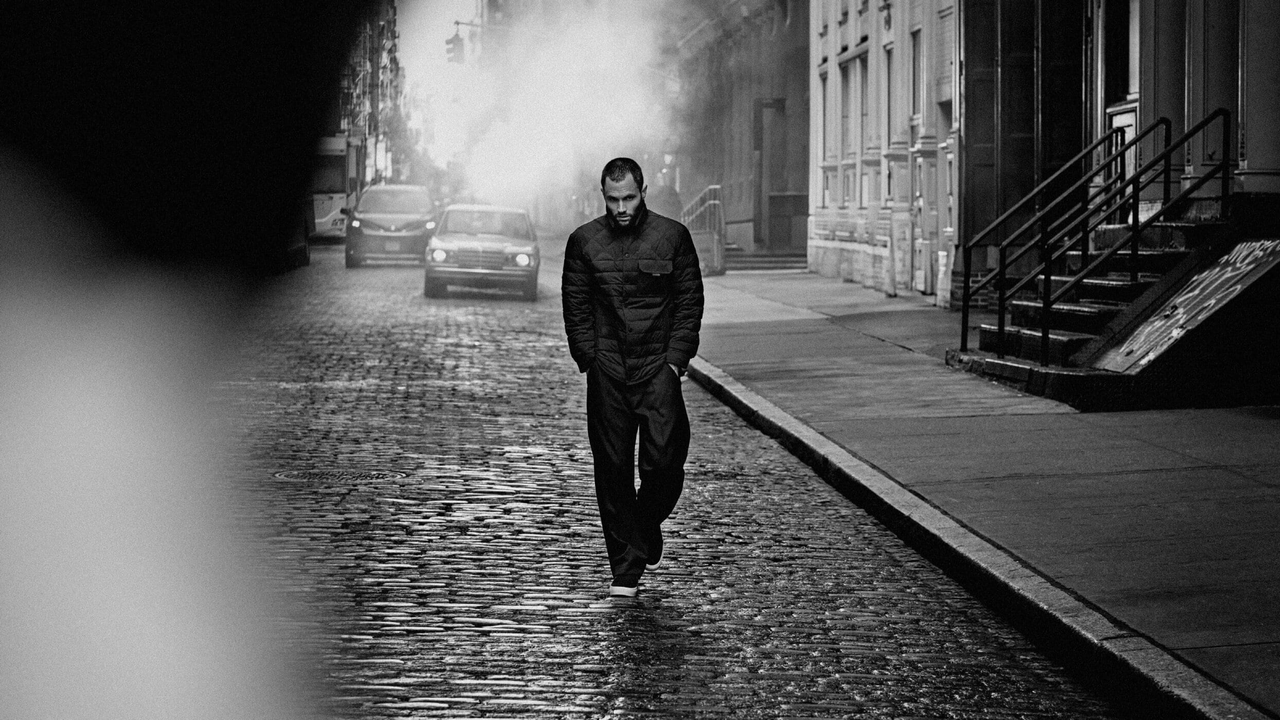 Man wearing a dark quilted puffer jacket walking down a cobblestone street with cars and buildings in the background.