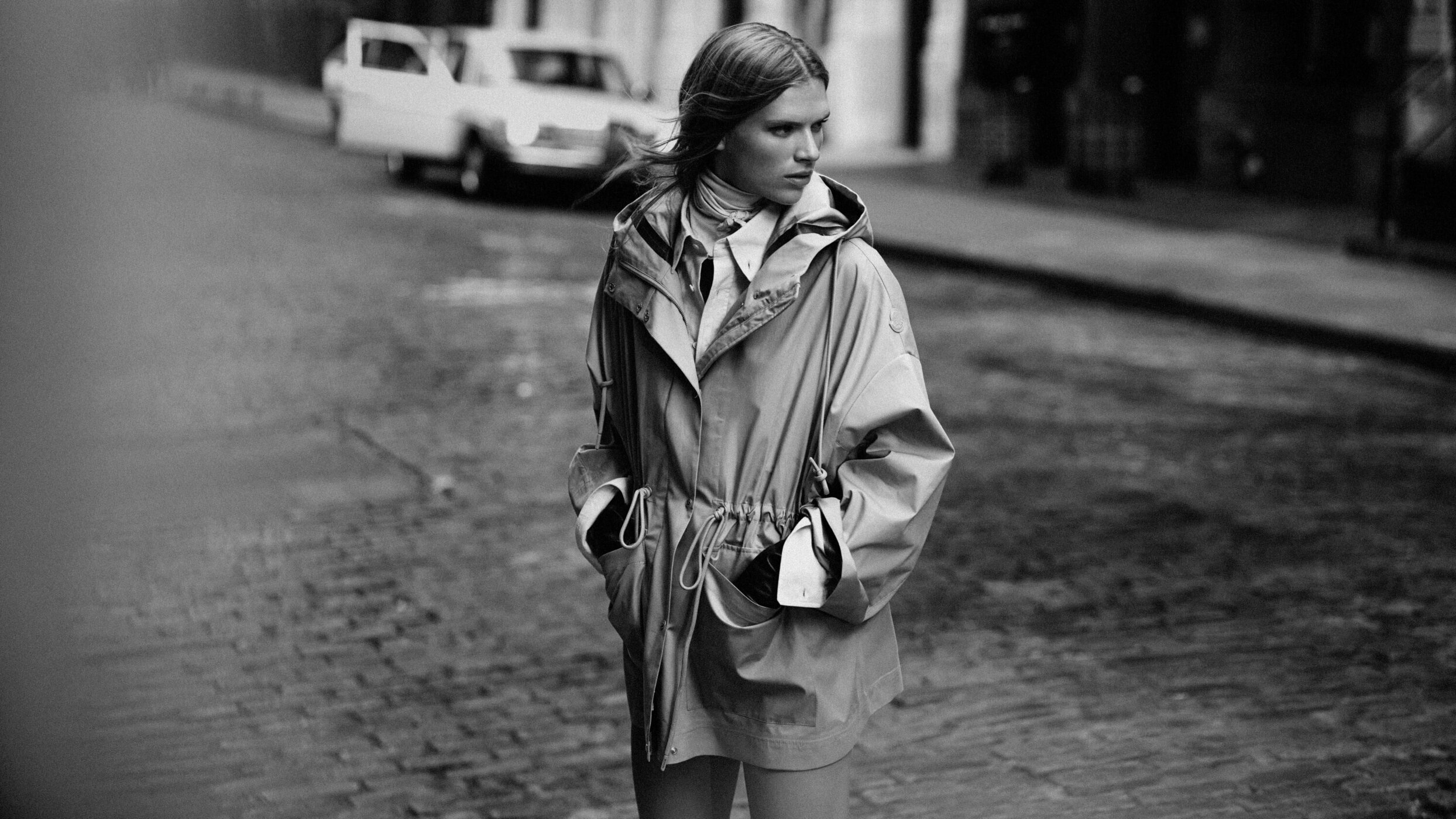 Monochromatic fashion photograph of a model in a hooded parka jacket standing on a cobblestone street with a car in the background.