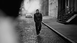 Man wearing a dark quilted puffer jacket walking down a cobblestone street with cars and buildings in the background.