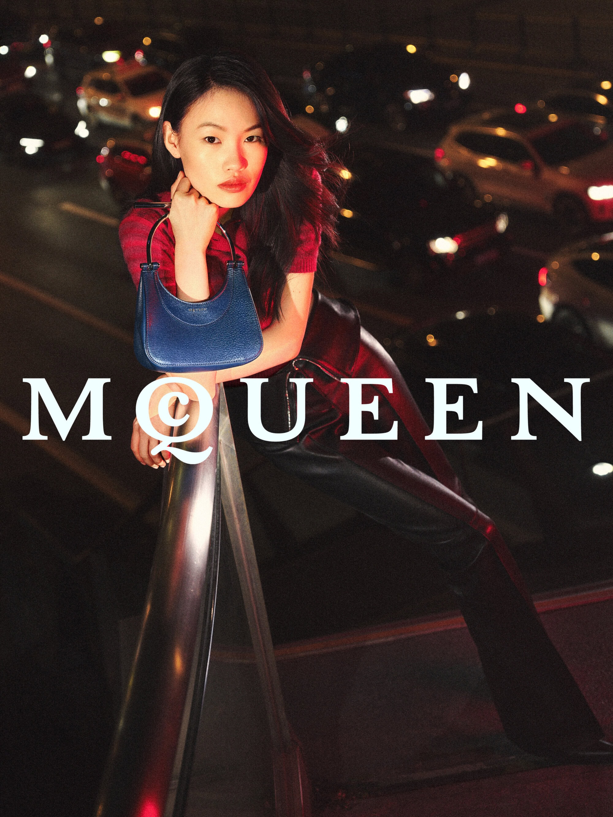 Model Zhang Youhao leaning on a railing holding a blue leather handbag against a blurred night street with car traffic.