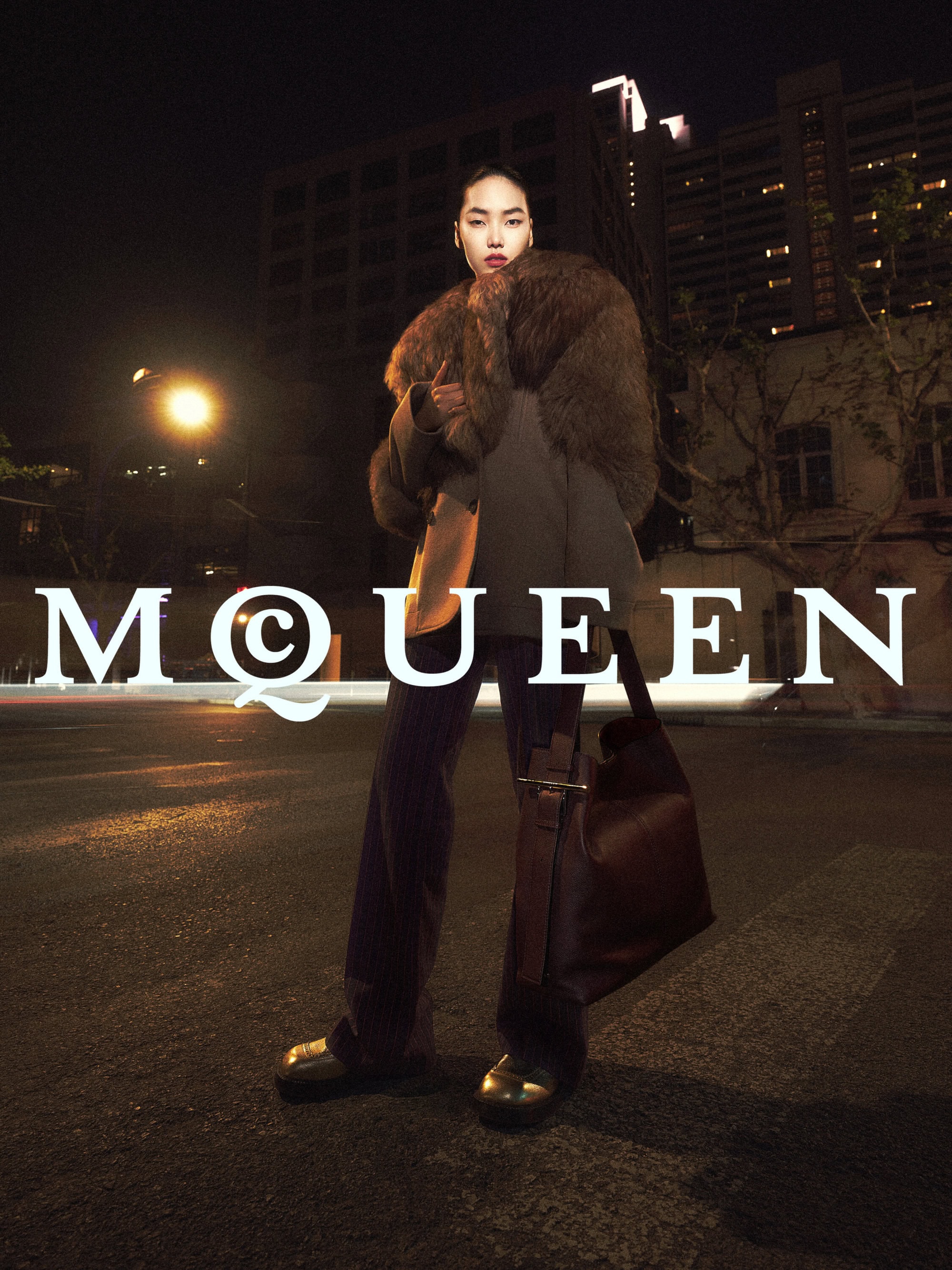 Model wearing a fur collar jacket and holding a large shoulder bag standing outdoors on a city street at night under warm street lighting