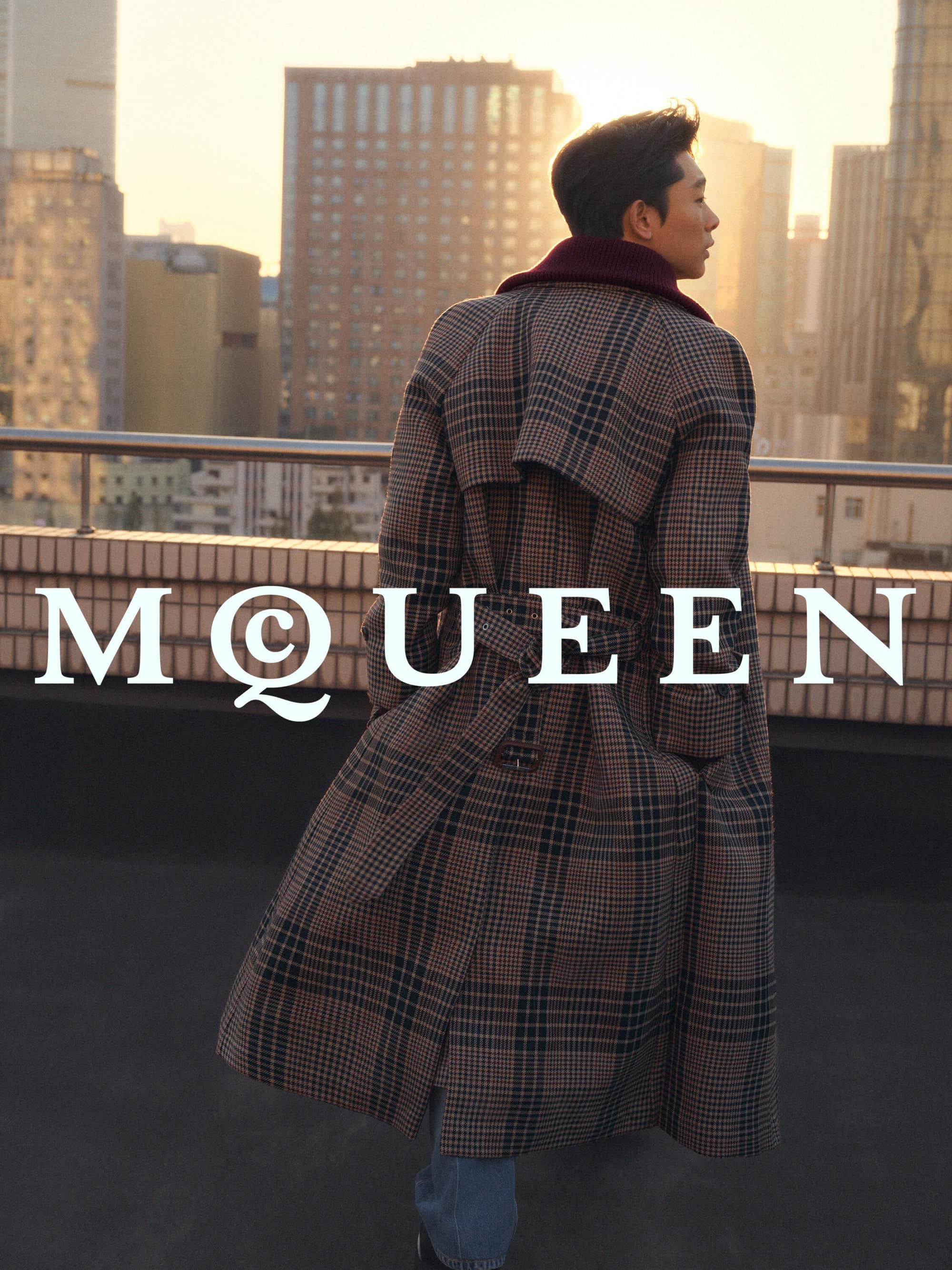 Man wearing a long plaid trench coat standing on a rooftop at sunset with city skyscrapers in the background