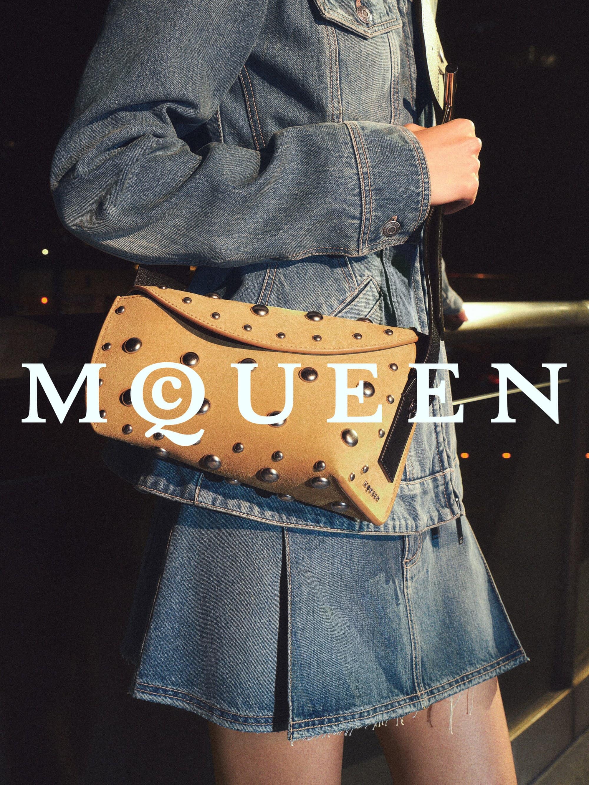 Model wearing a denim jacket and pleated denim skirt carrying a studded tan leather handbag in a night setting.