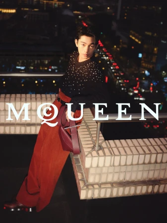 Model Zhang Youhao leaning on a rooftop railing at night wearing a dark embellished sweater and red trousers for McQueen
