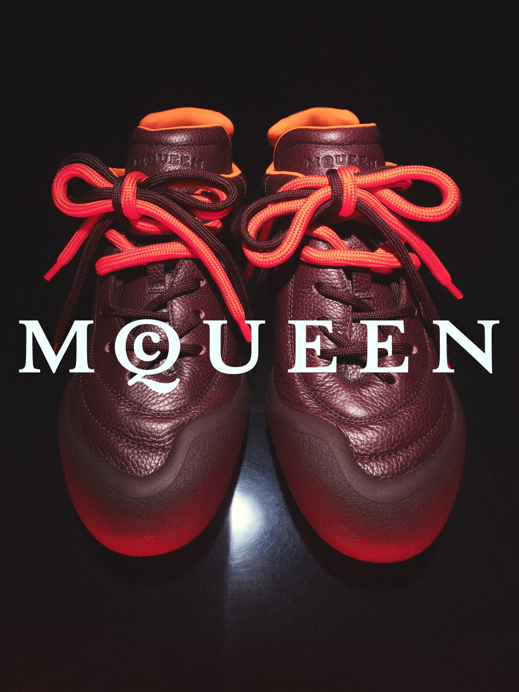 Pair of burgundy leather sneakers with bright orange laces displayed against a dark background with McQueen branding overlay.