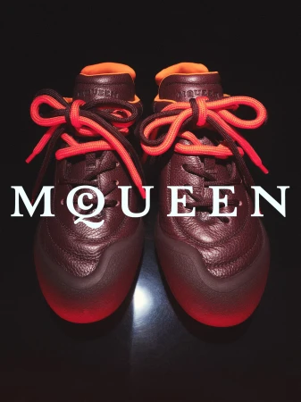 Pair of burgundy leather sneakers with bright orange laces displayed against a dark background with McQueen branding overlay.