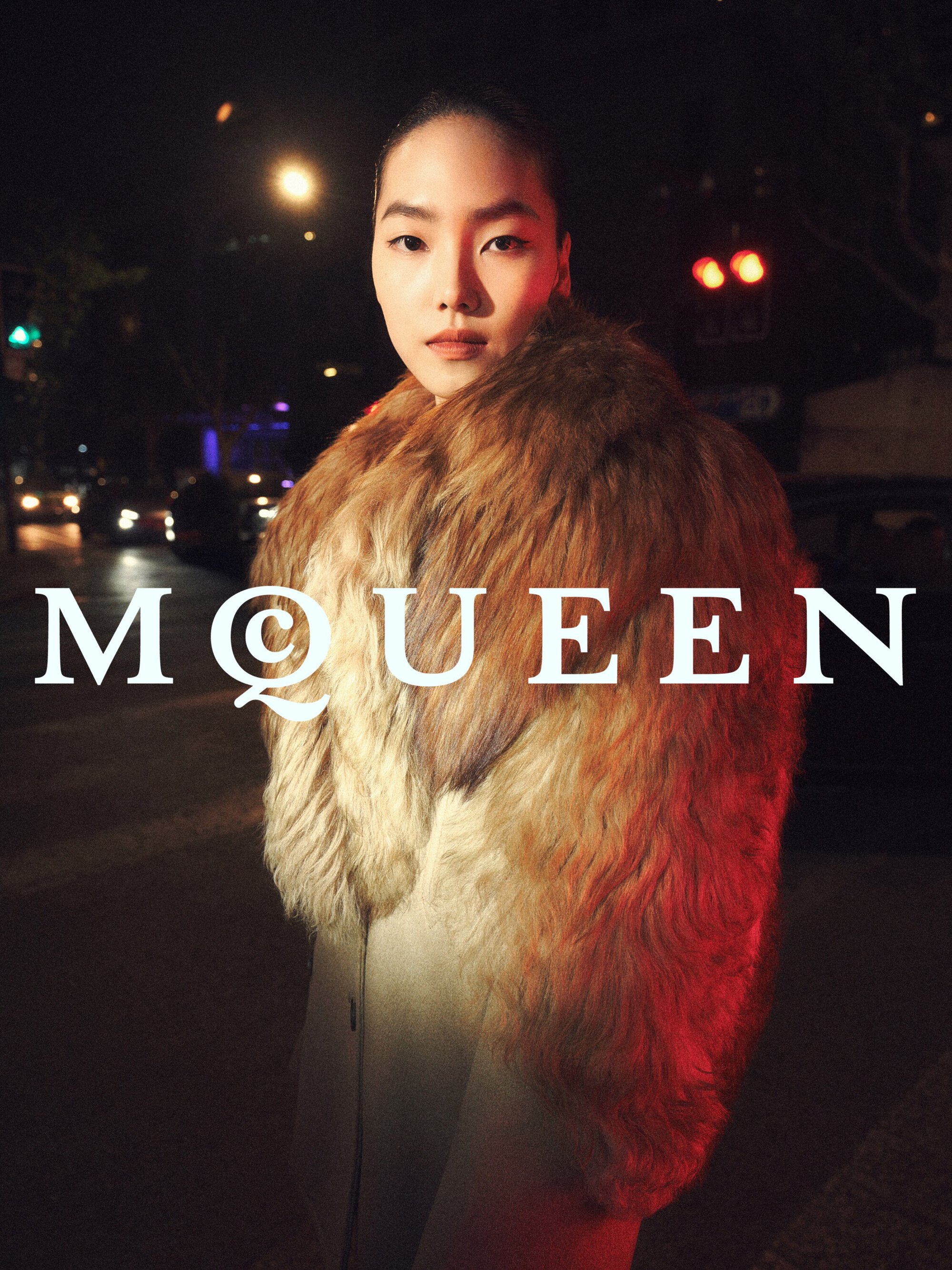 East Asian male model wearing a furry collar coat on a city street at night with red and white lighting.