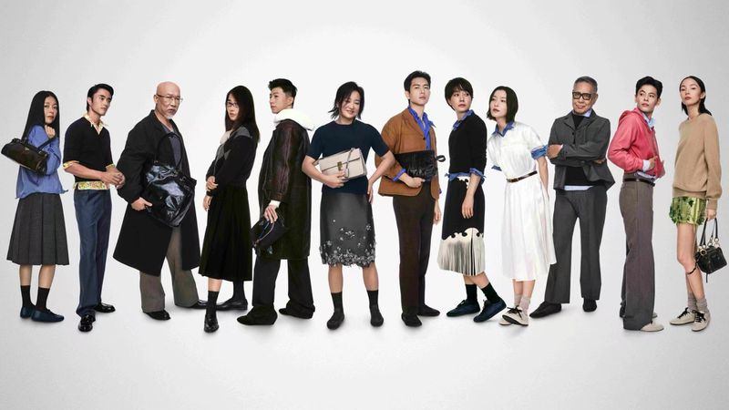 A group of models and creative figures standing in a line against a neutral studio background wearing Prada campaign apparel.