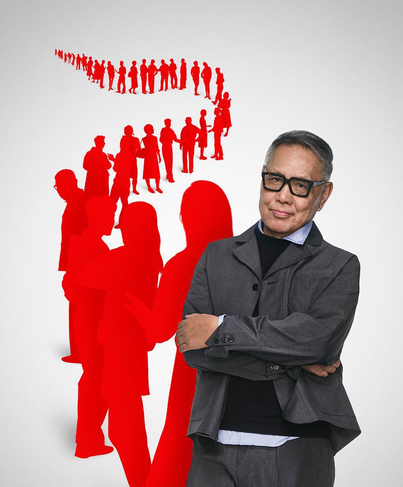 Elderly man wearing thick black-framed glasses and a grey blazer standing before a red silhouette graphic against white seamless background