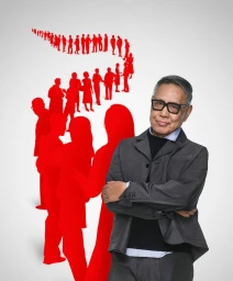 Elderly man wearing thick black-framed glasses and a grey blazer standing before a red silhouette graphic against white seamless background