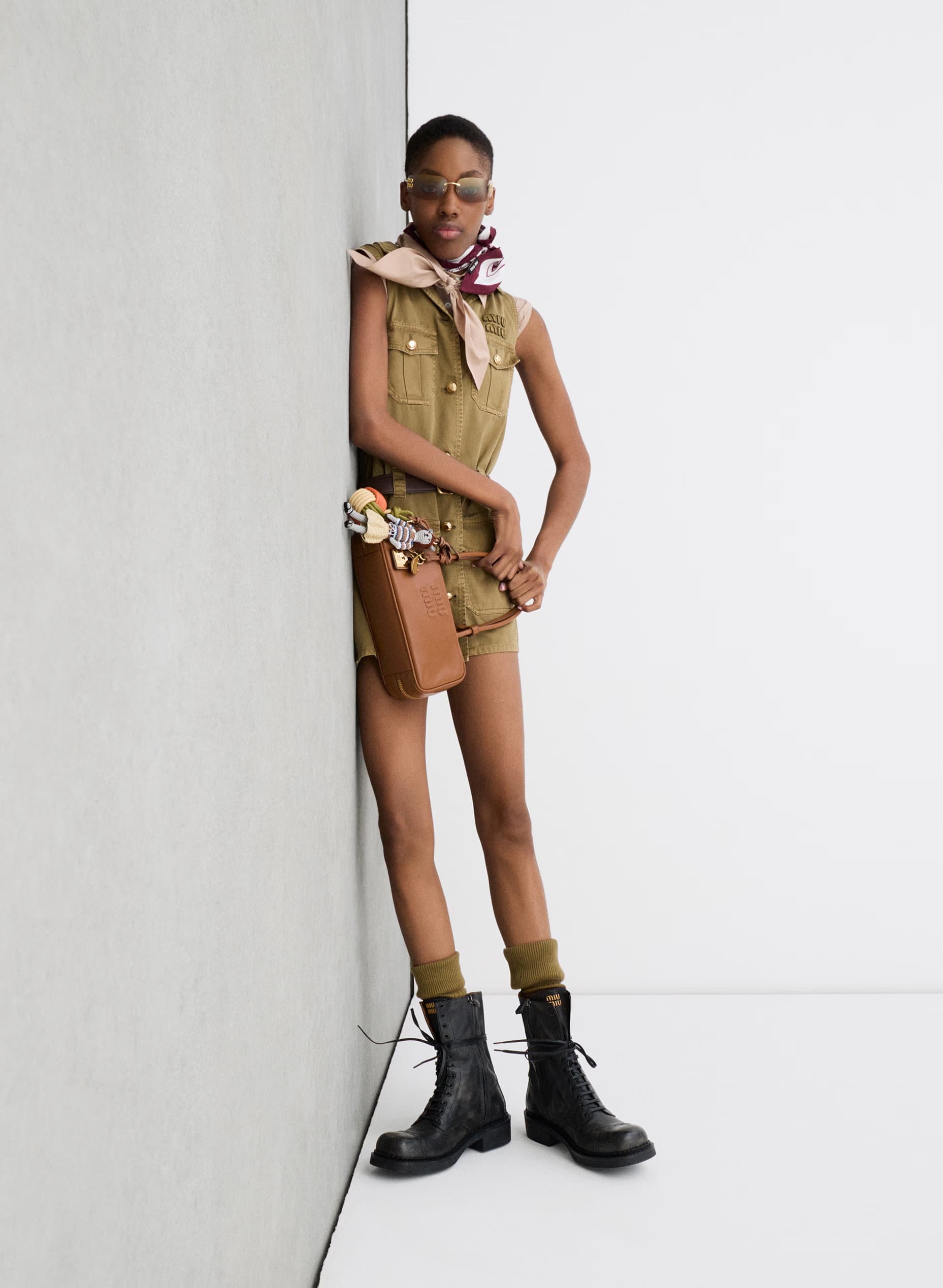 Model wearing utility romper, scarf, and boots leaning against a corner wall for Miu Miu Prelude 25 campaign.
