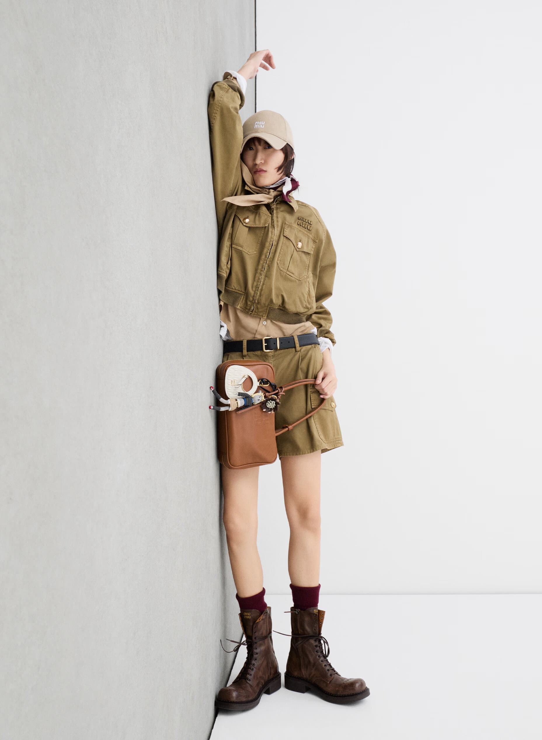 Model in olive utility jacket, shorts, and cap leaning against a white studio corner, holding a brown bag.