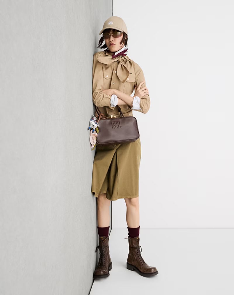 Model in khaki shirt and skirt wearing cap and dark sunglasses leaning against a grey and white wall holding brown handbag