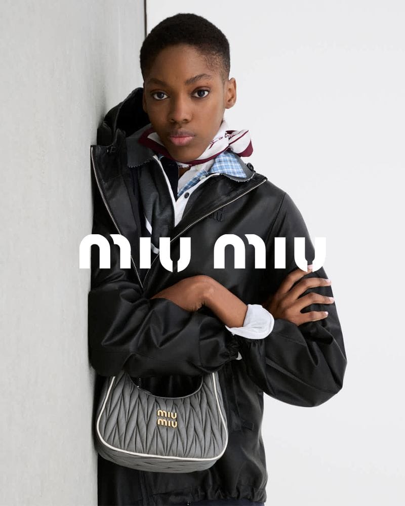 Model wearing a black jacket and holding a quilted grey handbag leaning against a plain wall for Miu Miu