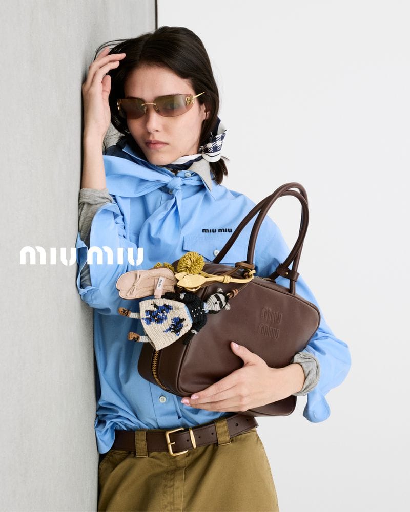 Model leaning against a wall wearing a light blue shirt, tinted sunglasses, and holding a brown leather handbag.
