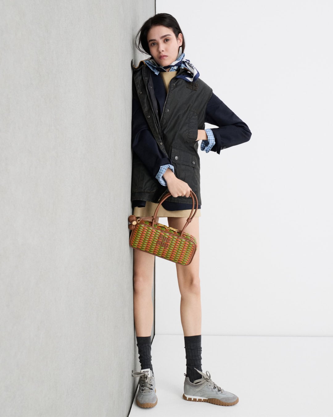 Model leaning against a grey wall wearing a dark vest, plaid shirt, and sneakers while holding a woven handbag.