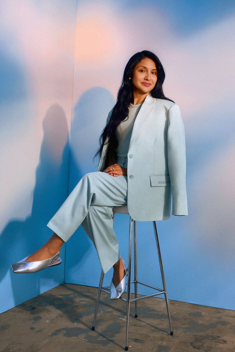 A stylish young woman with long dark hair is sitting on a stool wearing a light blue suit and silver flats in a studio setting.