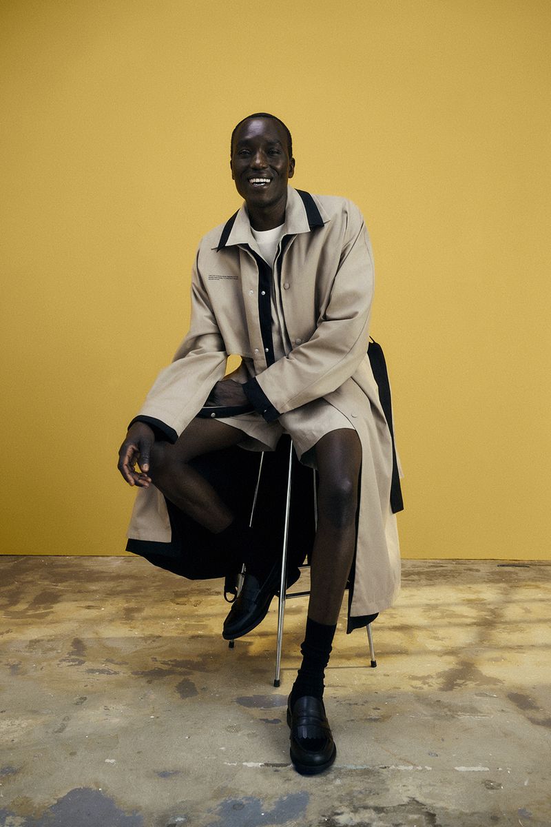 Full shot of a black male model wearing a light brown trench coat and black loafers while sitting on a stool in a studio.