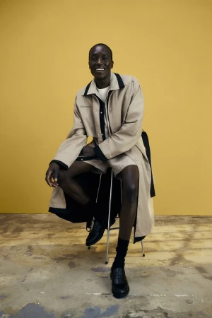 Full shot of a black male model wearing a light brown trench coat and black loafers while sitting on a stool in a studio.
