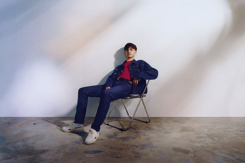 Young man in a denim jacket and jeans sitting on a metal chair in a photo studio, looking at the camera with a neutral expression.