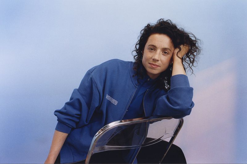 Portrait of a brunette woman in a blue zip-up jacket, leaning against a transparent chair, smiling gently in a softly lit interior space.