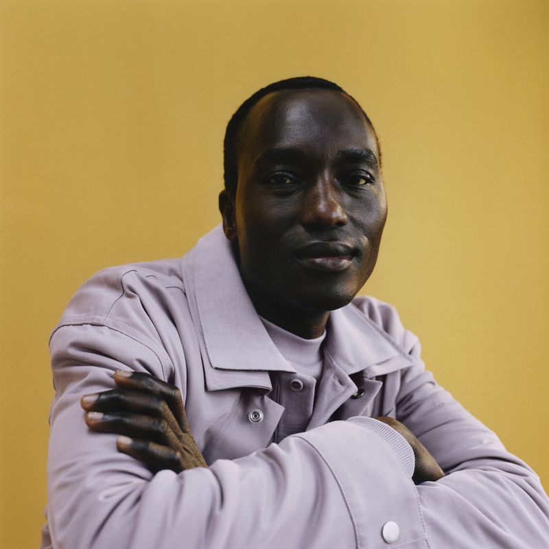Portrait of a dark-skinned man in a lavender jacket posing in front of a yellow backdrop with his arms crossed.