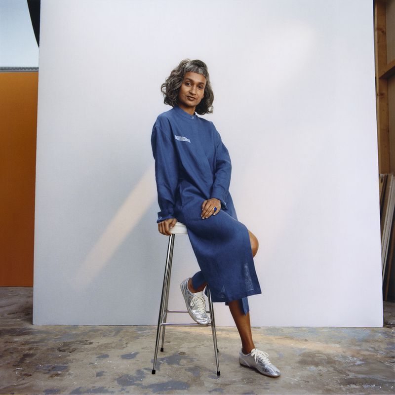 A woman sits on a stool in a blue dress and metallic sneakers against a white background with a concrete floor in a studio setting.