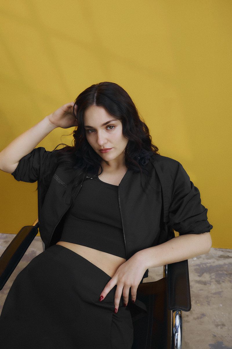 Brunette woman in a black top and jacket sits in a chair against a mustard-yellow background, striking a relaxed pose with her hand in her hair.