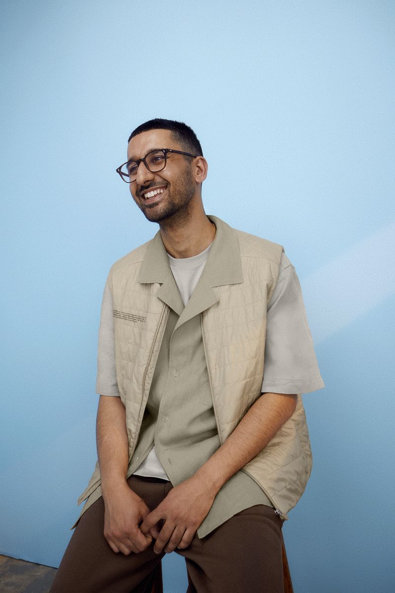 Smiling man in glasses wearing a tan vest and brown pants sitting against a light blue backdrop in a studio setting looking up.