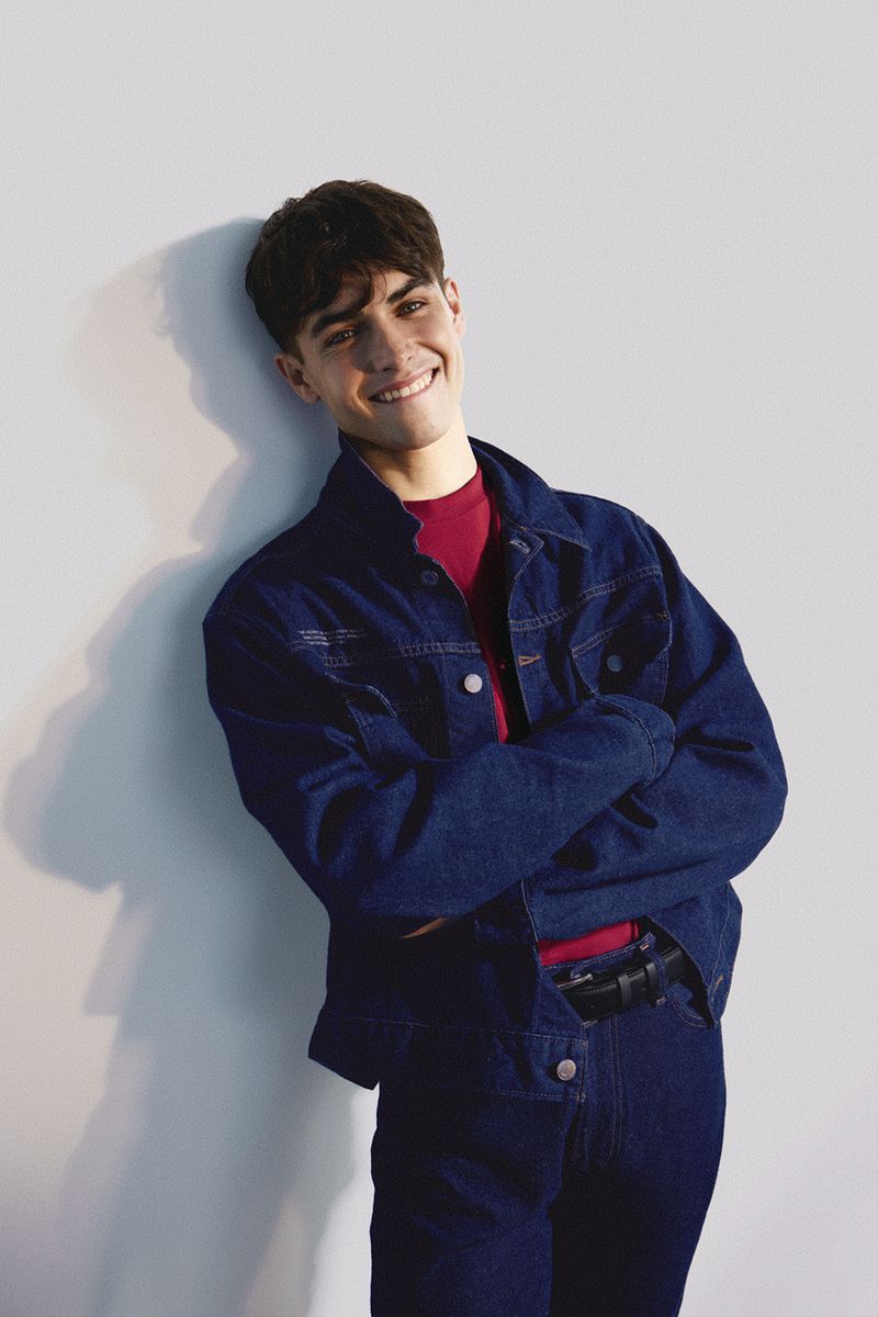 Smiling young adult male in dark denim jacket and jeans poses in front of a white wall with a casual pose in a photo studio.