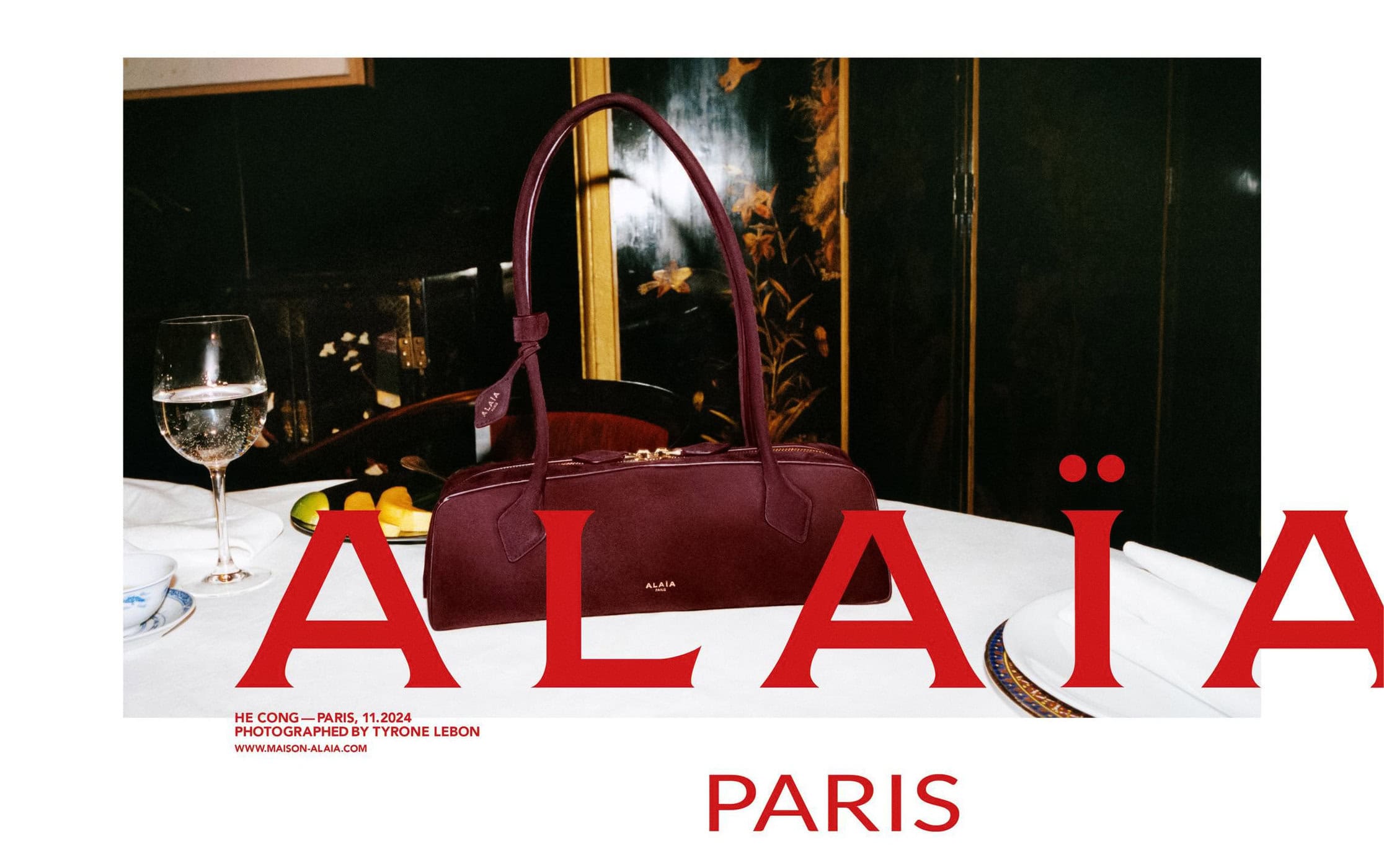 Burgundy leather shoulder bag resting on a set dining table next to a wine glass under dark paneling for Alaia