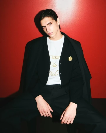 Model seated against a vibrant red wall wearing a black suit jacket, dark trousers, and white graphic t-shirt.