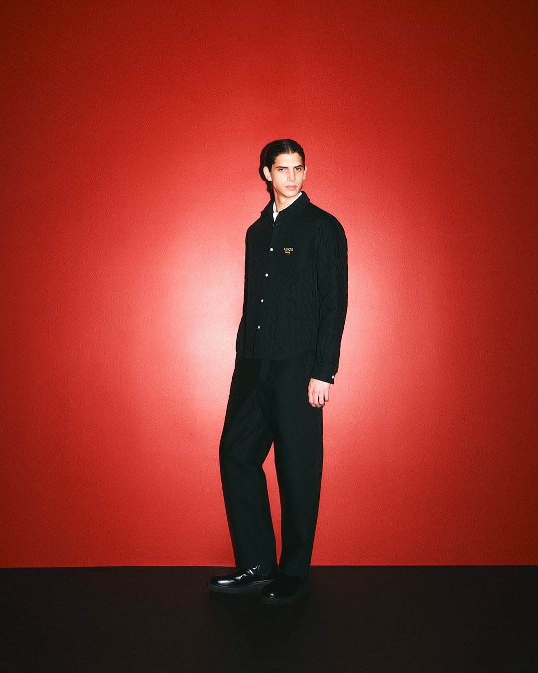 Model wearing a dark long-sleeved button-down shirt and trousers against a vibrant red backdrop for Kenzo.